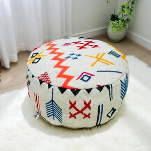 May include: Round, off-white pouf with a colorful geometric pattern. The design includes orange zigzags, red crosses, blue and yellow shapes, and black triangles. The pouf is placed on a white, fluffy rug, and is a decorative accent for any living space.