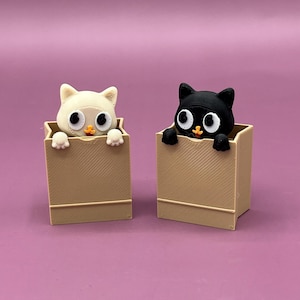 May include: Two cat figurines, one cream-colored and one black, peek out from tan boxes. The cats have large, round eyes and small orange beaks. The boxes have a simple, rectangular design with diagonal lines on the front.