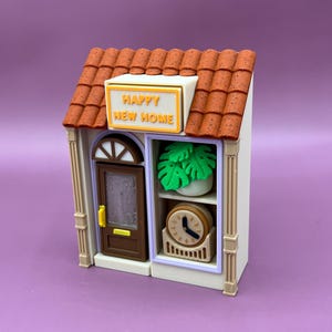 以下が含まれることがあります： 茶色の瓦屋根と「HAPPY NEW HOME」と書かれた看板が付いたミニチュアハウスモデル。家には茶色のドア、植物と時計のある窓、ベージュのトリムがあります。全体的なデザインは気まぐれで装飾的です。
