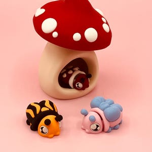 Puede incluir: Una caprichosa figura en forma de hongo con un sombrero rojo y lunares blancos, que alberga una mariquita. Dos figuras de insectos adicionales, una naranja y negra, la otra rosa y azul, se exhiben sobre una superficie rosa. Probablemente sean artículos decorativos.