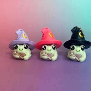 May include: Three whimsical frog figurines, each wearing a different colored wizard hat with a crescent moon accent. The hats are lavender, hot pink, and black. The frogs are light green with large black eyes and hold a tan-colored staff.