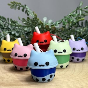 May include: Six colourful cat-shaped boba tea cups. Each cup is a different colour: yellow, pink, red, light blue, green, and purple. The cups have black dot details and a white straw.
