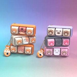 Op de afbeelding: Twee stapels van drie rechthoekige houders voor mechanische toetsenbordtoetsen in oranje, wit, grijs en lavendel. Elke houder bevat drie toetsen met kattengezichten en pootafdrukken. Een kleine oranje visvormige bedel hangt aan een gouden kogelketting.