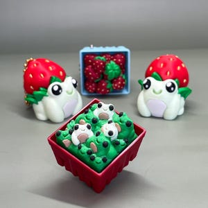 May include: A collection of whimsical frog-themed miniatures. Two white frogs with strawberry hats and green leaves sit on either side of a red container filled with frog-shaped figures in a green base. A blue container holds miniature strawberries.
