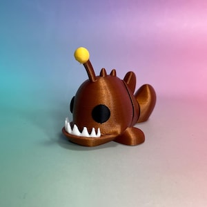 May include: A small, 3D-printed anglerfish figurine in a metallic bronze finish. The creature features a prominent yellow lure extending from its head, large black circular eyes, and a wide mouth lined with sharp, white, triangular teeth.