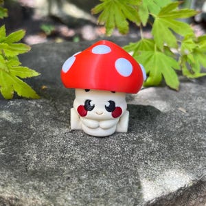 Puede incluir: Una caprichosa figura de hongo con un sombrero rojo con lunares blancos. El hongo tiene una cara sonriente con ojos negros y mejillas rojas. La figura es blanca y tiene un diseño lindo y de dibujos animados.