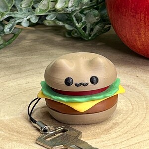 Peut inclure: Un porte-clés fantaisie représentant un mini-burger avec un visage de chat. Le burger a un pain beige, de la laitue verte, du fromage jaune et une tomate rouge. Le porte-clés est attaché à une clé argentée.