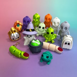 May include: A collection of Halloween-themed miniature figurines in various colors. The assortment includes ghosts, aliens, pumpkins, a witch's cauldron, a finger, and a frog. The figurines are arranged on a gradient purple and pink background.