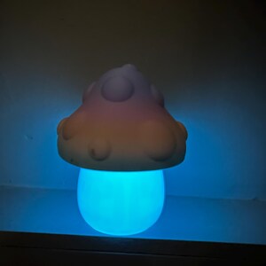 Może przedstawiać: Nocna lampka w kształcie grzyba w pastelowych kolorach z niebieskim blaskiem. Grzyb ma zaokrąglony wierzchołek z białymi kropkami i gładką, zaokrągloną podstawę.
