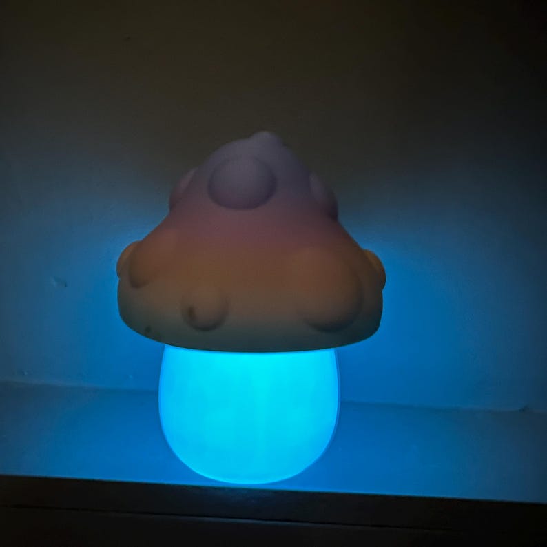 Może przedstawiać: Nocna lampka w kształcie grzyba w pastelowych kolorach z niebieskim blaskiem. Grzyb ma zaokrąglony wierzchołek z białymi kropkami i gładką, zaokrągloną podstawę.