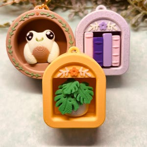 May include: Three miniature decorative pendants. One features a frog, another a bookshelf with books, and the third a potted plant. The frog is in a brown circular frame with green leaves. The bookshelf is in a lilac frame. The plant is in an orange frame.
