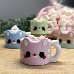 May include: A selection of pastel-coloured cat-shaped mugs, each filled with marshmallows. The mugs have white handles and facial features. A bag of marshmallows is visible in the background.