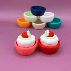 May include: A collection of colorful, small, scalloped-edge bowls in various colors, including blue, green, white, orange, and pink. Two miniature desserts with strawberry toppings are in the foreground. The background is a solid purple.