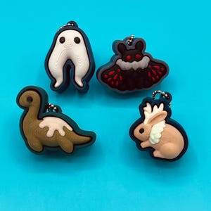 Può includere: Quattro portachiavi colorati con disegni di cartoni animati. I portachiavi includono un fantasma bianco, una falena nera con accenti rossi, un dinosauro marrone con una macchia bianca e un coniglio marrone con pelliccia bianca. Ogni portachiavi ha una catena argentata e un bordo blu.