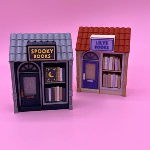May include: Two miniature bookshop models. One shop has a gray roof and a sign that reads "Spooky Books." The other shop has a red roof and a sign that reads "Lilys Books." Both shops have doors and shelves filled with books.