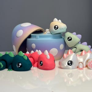Op de afbeelding: Een verzameling kleurrijke, 3D-geprinte dinosaurusfiguren in verschillende kleuren, waaronder teal, roze en iriserend wit. Sommige dinosaurussen staan in de buurt van een paddenstoelvormige container met een pastel kleurenschema. De figuren hebben grote, ronde ogen.