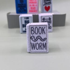 Pode incluir: Uma coleção de pequenos amuletos retangulares em forma de livro. O amuleto central diz "BOOK WORM" com um gráfico de verme. Outros amuletos apresentam texto e gráficos, incluindo um coração e as palavras "I Like Big Books".