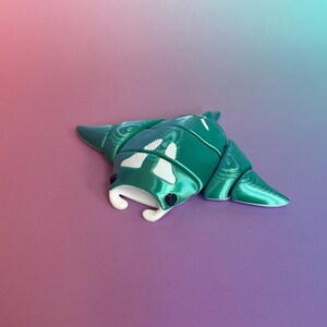 May include: A teal and white 3D-printed articulated bug toy. The segmented body has white markings and a curved, white mouth. The toy is set against a pink and blue gradient background.