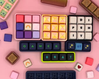May include: An overhead shot displays colorful, square-shaped items on a pink surface. There are several trays with square compartments filled with various colors, including pink, purple, yellow, and white. Some trays have letters and numbers.