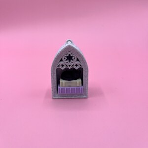 以下が含まれることがあります： 黒猫が入った、グレーのゴシック様式のミニチュアキャットハウス。家には花の模様のアーチ型のトップがあり、猫と本の山を置く小さな棚があります。本はベージュと紫です。
