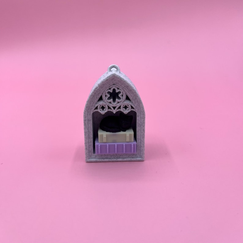 以下が含まれることがあります： 黒猫が入った、グレーのゴシック様式のミニチュアキャットハウス。家には花の模様のアーチ型のトップがあり、猫と本の山を置く小さな棚があります。本はベージュと紫です。
