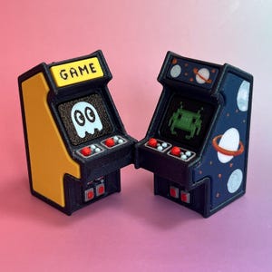 Könnte beinhalten: Zwei Miniatur-Spielautomaten mit schwarzen Rahmen. Das linke Gerät ist gelb mit dem Wort GAME oben und einer weißen Geistergrafik auf dem Bildschirm. Das rechte Gerät ist dunkelblau mit Weltraummotiv und einer grünen Alien-Grafik auf dem Bildschirm.