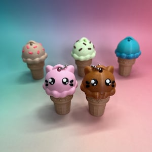 Può includere: Una collezione di cinque ciondoli a forma di cono gelato. I ciondoli presentano una varietà di colori, tra cui rosa, marrone, verde e blu. Ogni ciondolo ha una faccia di gatto o orso sulla parte superiore del cono. I coni sono di colore marrone chiaro.