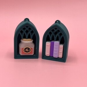以下が含まれることがあります： 2つの濃いティール色のミニチュアアーチ型棚。1つの棚には、コルクの蓋と三日月と星のラベルが付いた小さな瓶が入っています。もう1つの棚には、ピンク、紫、ラベンダーの3冊の小さな本が入っています。