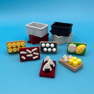 Könnte beinhalten: Eine Sammlung miniaturisierter japanischer Lebensmittel, darunter Sushi und Reisbällchen, auf einer blauen Oberfläche angeordnet. Die Lebensmittel sind in verschiedenen Farben wie Weiß, Gelb, Rot und Braun gehalten. Es gibt auch kleine, rechteckige Behälter.