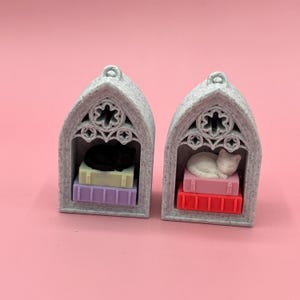 以下が含まれることがあります： ミニチュアの灰色のゴシックアーチが2つあり、それぞれに眠っている猫の置物があります。1匹の猫は黒、もう1匹は白です。どちらの猫もカラフルな本の山の上にいます。背景は無地のピンク色です。