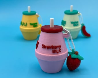 Puede incluir: Tres colgantes en miniatura con forma de botellas de leche. El colgante delantero es rosa con el texto "Strawberry Milk". Un colgante de fresa roja cuelga del lateral. Otros colgantes son amarillos y verdes. Todos tienen pajitas blancas y están sobre un fondo azul.