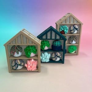 Pode incluir: Miniaturas de prateleiras de estufa de madeira exibindo esculturas artesanais de suculentas e cristais. As prateleiras são bege e verde-azulado, com várias plantas e cristais coloridos. O fundo tem um gradiente de rosa, azul e roxo.
