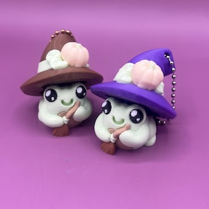 May include: Two whimsical, handmade clay figurines. Each figure is a light green blob with large black eyes, a small smile, and a tiny broom. They wear witch hats, one brown and one purple, adorned with a pink pumpkin and white accents.