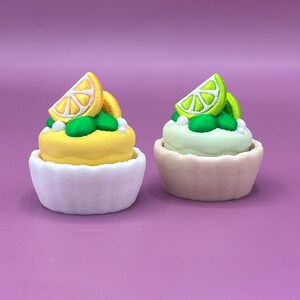 以下が含まれることがあります： 2つのミニチュア装飾カップケーキ。1つはレモンスライスが上に乗った黄色、もう1つはライムスライスが乗った薄緑色です。どちらも白と薄ピンクのカップケーキホルダーに入っています。緑と白のアクセントがデザインを完成させ、紫色の背景に配置されています。