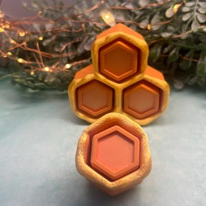 May include: Orange and yellow honeycomb-shaped decorative objects. The objects have a hexagonal design, with a textured surface. The objects are arranged on a light blue surface, with greenery and fairy lights in the background.