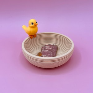 Puede incluir: Un pequeño cuenco beige y acanalado que contiene varias monedas, con una figura de pollito amarillo posada en el borde. El pollito tiene patas naranjas y un ojo negro. El fondo es de color lila claro.
