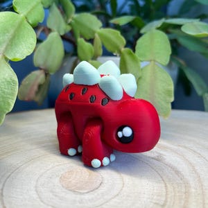 Może przedstawiać: Mały, czerwony, wydrukowany w 3D dinozaur z białymi kolcami i czarnymi oczami. Dinozaur stoi na drewnianej powierzchni.