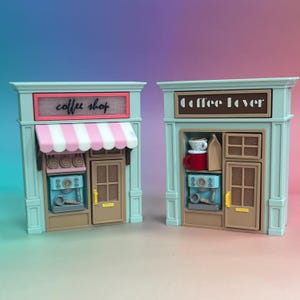 Può includere: Due modelli di vetrine in miniatura color azzurro con porte marroni e maniglie gialle. Il negozio a sinistra ha una tenda a righe rosa e bianche e un'insegna con scritto coffee shop. Quello a destra ha un'insegna coffee lover con una tazza rossa.