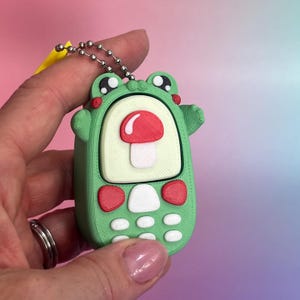 May include: A light green 3D printed keyring shaped like a frog with a screen displaying a red and white mushroom icon. The device features a light green body, two red oval buttons, and six small white buttons arranged in a grid below the screen.
