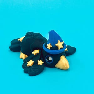 Puede incluir: Una caprichosa figura de cuervo verde oscuro con pico y patas amarillas, que lleva un sombrero de mago azul adornado con estrellas doradas. El cuervo está tumbado sobre un fondo turquesa liso, creando una estética lúdica y mágica.