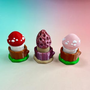 以下が含まれることがあります： 一列に並んだ3つの小さなキノコのフィギュア。左のキノコは茶色の切り株の上にあり、赤地に白い斑点があります。中央のキノコは紫色のベースの上にあり、表面に凹凸があります。右のキノコは茶色の切り株の上にあり、薄いピンク地に白い斑点があります。