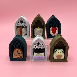 May include: Seven gothic-style pendants in different colours. Each pendant has a miniature scene: a dragon with books, candles, a heart, a clock, a cat, and a frog. The pendants are set against a pink background.