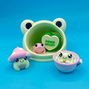 以下が含まれることがあります： カエルをテーマにした、気まぐれな置物とアクセサリーのコレクション。明るい緑色のカエル型ボウル、ピンクのキノコフィギュア、ティーカップに入ったカエル、そして「FROG CRAZY」の文字が入ったハート型のキーチェーンが含まれています。明るい青色の背景に配置されています。