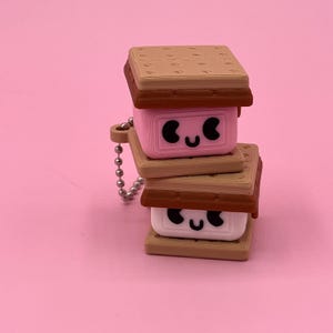 Puede incluir: Un dije apilado que se asemeja a un s'more, con galletas Graham, chocolate y una malvavisco sonriente. El dije está unido a una cadena plateada, sobre un fondo rosa.