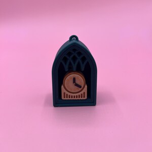以下が含まれることがあります： ゴシックアーチ型のフレームに入った、ティール色のミニチュア時計。 時計の文字盤はタン色で、黒い針と数字が付いています。 フレームは装飾的な透かし彫りのデザインで、上部に小さなループが付いています。 背景はピンク色です。