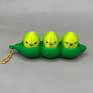 May include: A green pea pod keychain with three smiling yellow peas inside. The peas have black eyes and are nestled in a green pod. The keychain has a gold-colored chain.