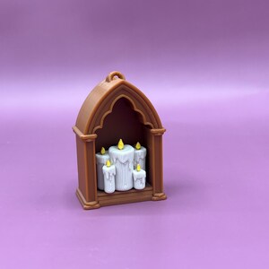 以下が含まれることがあります： 茶色のミニチュアゴシック様式のキャンドルホルダー。5本の白いろうそくがあり、ろうそくには黄色の炎が付いています。ホルダーはアーチ型のトップと、吊り下げ用の小さなループが付いています。