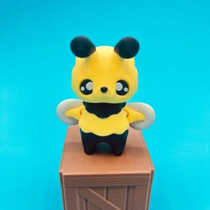 Könnte beinhalten: Eine 3D-gedruckte gelb-schwarze Bienenfigur mit großen, ausdrucksstarken Augen und kleinen Flügeln. Die Biene steht auf einer braunen Holzkiste vor einem hellblauen Hintergrund. Die Biene hat ein niedliches, cartoonhaftes Design.