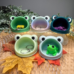 Puede incluir: Cinco cuencos con forma de rana en varios tonos de verde y azul. Cada cuenco contiene objetos pequeños como anillos, clips y una pequeña figura de rana. Los cuencos están colocados sobre hojas de otoño.