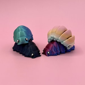 Könnte beinhalten: Zwei 3D-gedruckte, farbenfrohe Asselfiguren. Eine ist türkis und marineblau, die andere ein Farbverlauf aus Lila, Blau und Gelb. Jede hat einen segmentierten Körper und kleine, weiße Augen. Der Hintergrund ist einfarbig rosa.